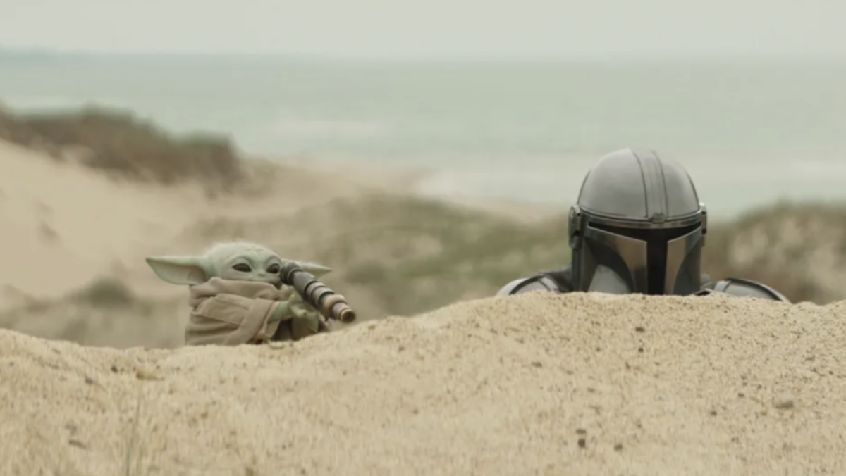 The Mandalorian and Grogu hiding behind a dune of sand.